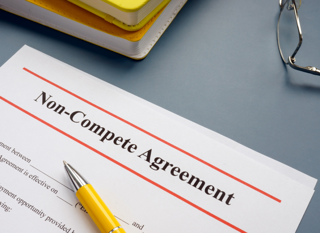 A printed non-compete agreement on a table near a stack of books with a pen on top of the contract.