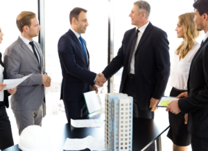 A business meeting of real estate investors takes place in an office setting. The real estate investors stand around a table with a building model on top. Two of the investors are shaking hands.