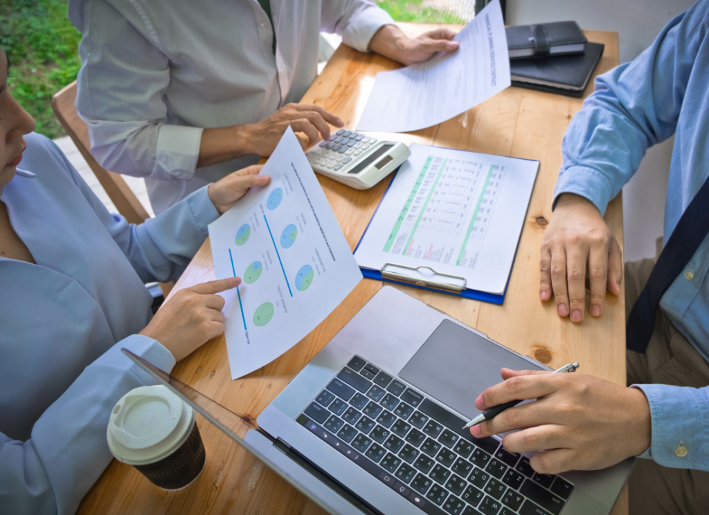 Three people are working at a desk with a laptop computer and reviewing paper documents.