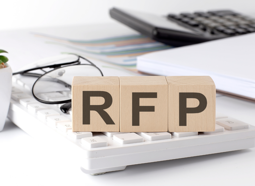 Three wood blocks with the letters RFP, or request for proposal, are on top of a keyboard on a white desk. There is a pair of glasses, a calculator, and a few loose documents in the background.