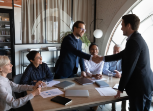 A group of HR professionals gathered at a table in an office working on outsourced HR tasks. Two individual in the group are standing and shaking hands across the table.