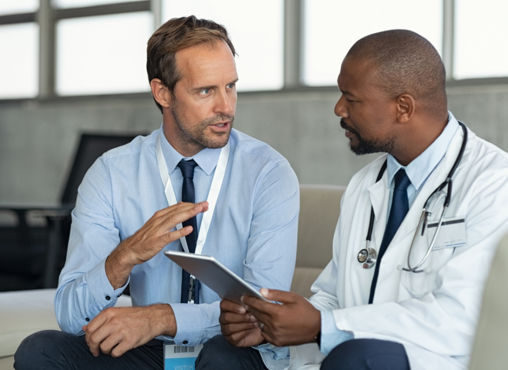 A male physician is speaking with an advisor about selling his medical practice. The physician is holding a tablet computer and speaking to the advisor sitting next to him.