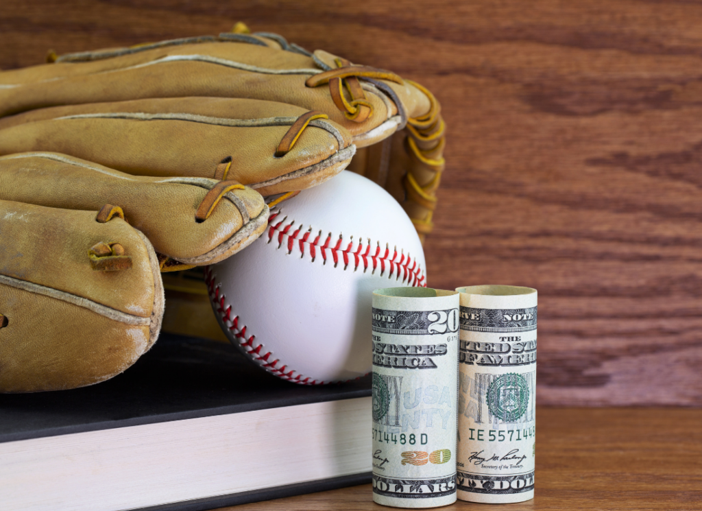 A text book, baseball, leather glove, and dollars are shown on a wood background, symbolizing student athlete compensation.