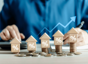 Wooden house models with percentage symbols sit on stacks of coins, representing real estate investment growth. In the background, a real estate investor is assessing the increasing value of the real estate investments.