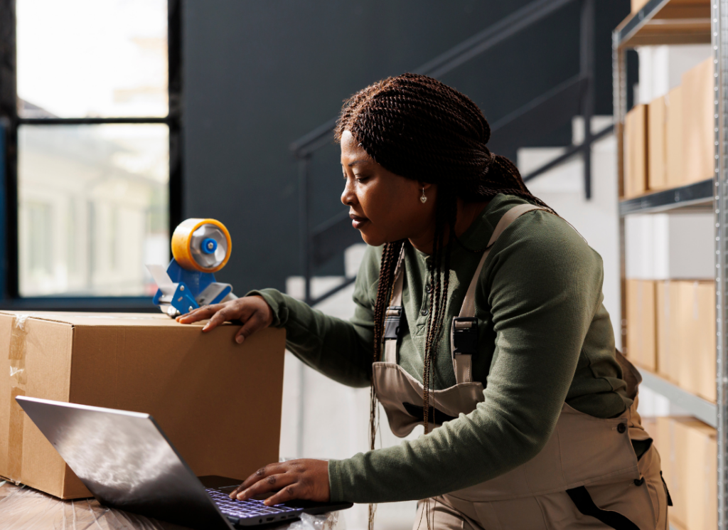 A warehouse worker in overalls manages inventory, using a laptop and handling a package, illustrating best practices in supply chain management.