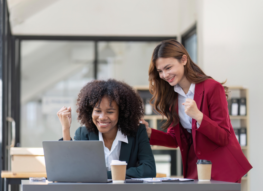 Two women in business attire smile and celebrate while looking at a laptop in an office setting, with coffee cups and paperwork on the table.