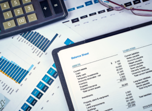 A balance sheet on a clipboard is surrounded by financial charts, a calculator, and glasses, illustrating financial statement analysis for manufacturers.