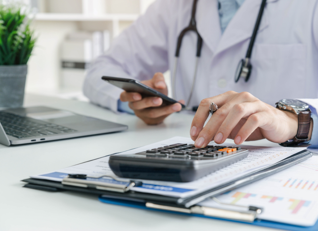 A doctor in a white coat with a stethoscope uses a calculator and smartphone at a desk with financial documents, highlighting accounting challenges in concierge medicine.