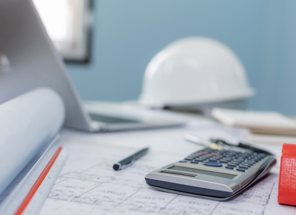A desk with construction documents, a calculator, pen, and rolled blueprints, with a white hard hat and laptop in the background, symbolizing financial support for construction bonding needs.
