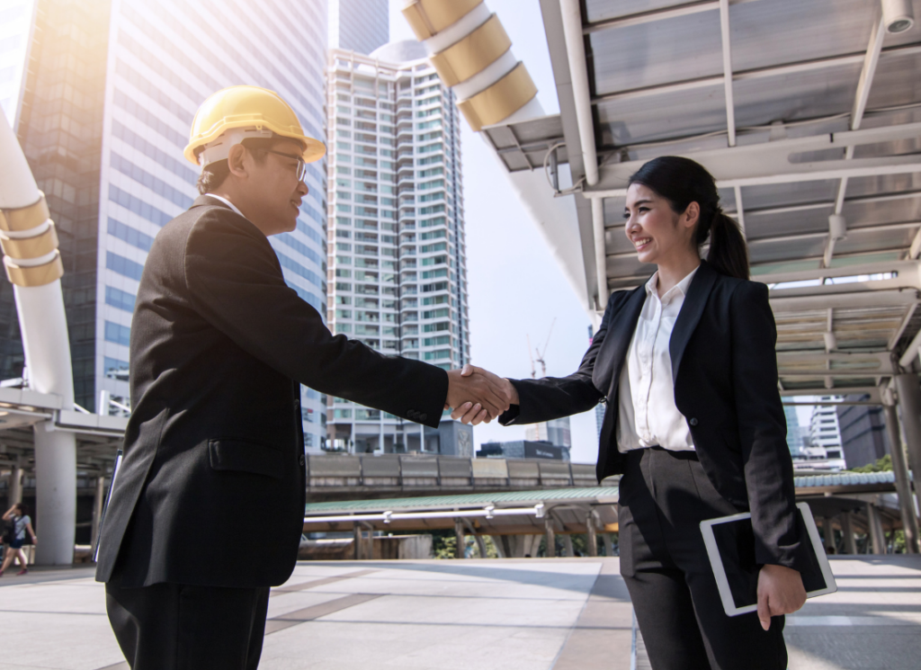 A construction professional wearing a hard hat and business suit shakes hands with a fractional construction CFO holding a tablet with tall buildings in the background.