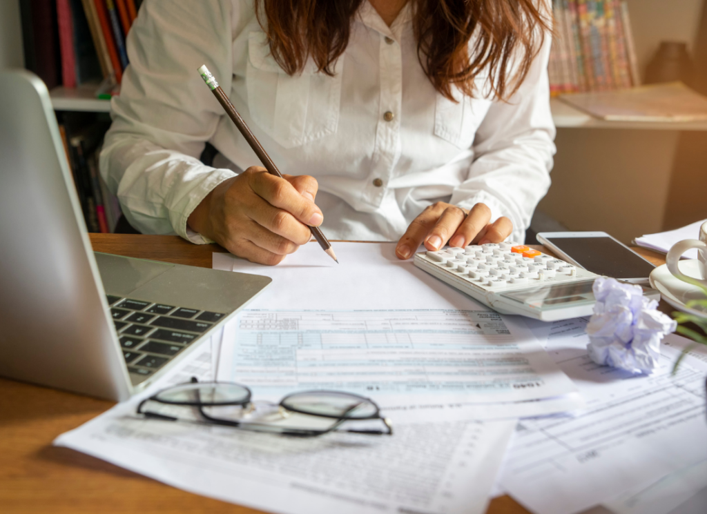 A person sits at a desk with tax forms, a calculator, and a laptop, reviewing documents and taking notes for tax planning.