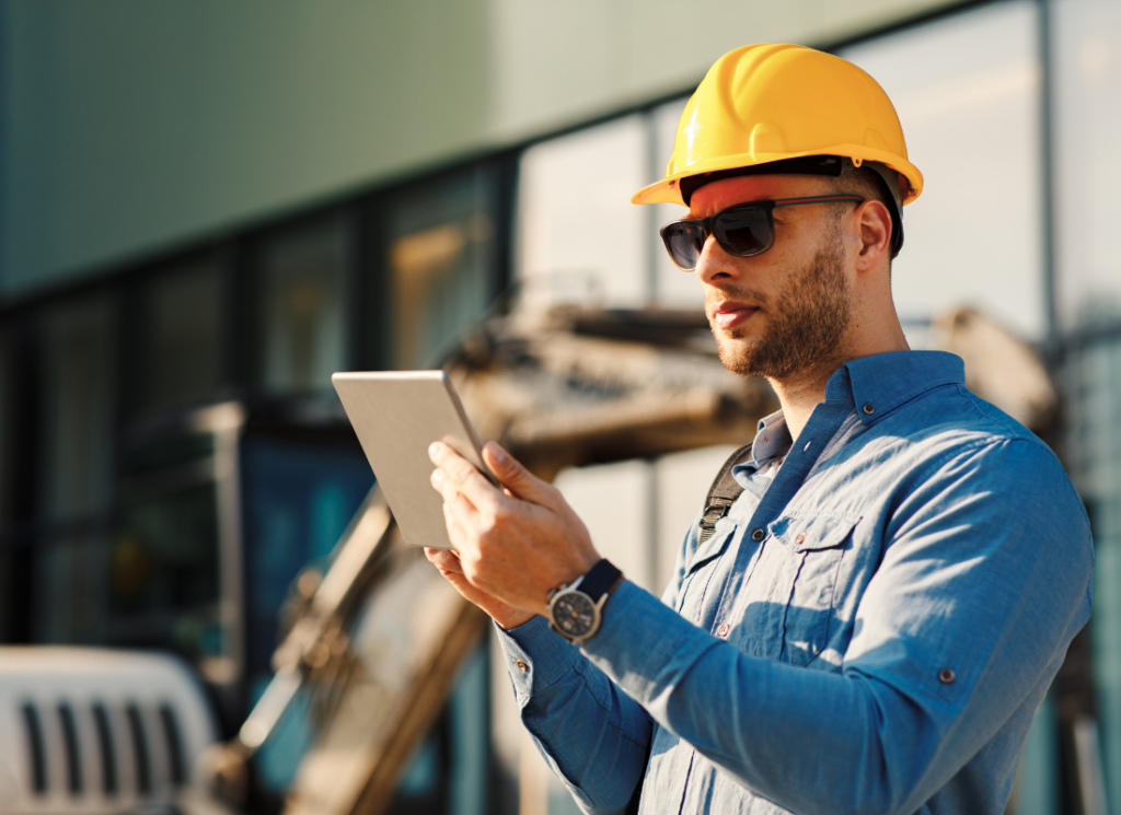 A construction professional wearing a hard hat and sunglasses uses a tablet at a job site.