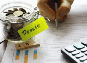 A glass jar labeled “Donate” filled with coins sits beside tax documents, a hand holding a pen, wooden letter tiles spelling “tax,” and a calculator—symbolizing nonprofit accounting.
