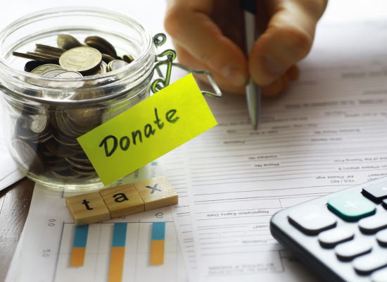 A glass jar labeled “Donate” filled with coins sits beside tax documents, a hand holding a pen, wooden letter tiles spelling “tax,” and a calculator—symbolizing nonprofit accounting.