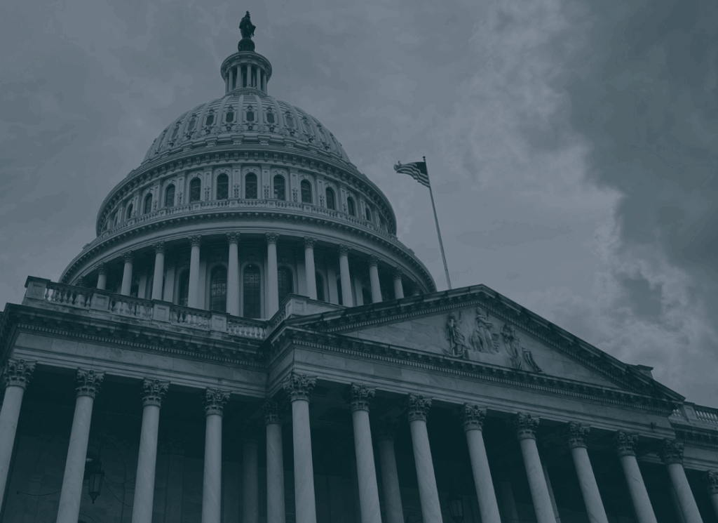 The U.S. Capitol building is shown under a cloudy sky with the American flag waving.