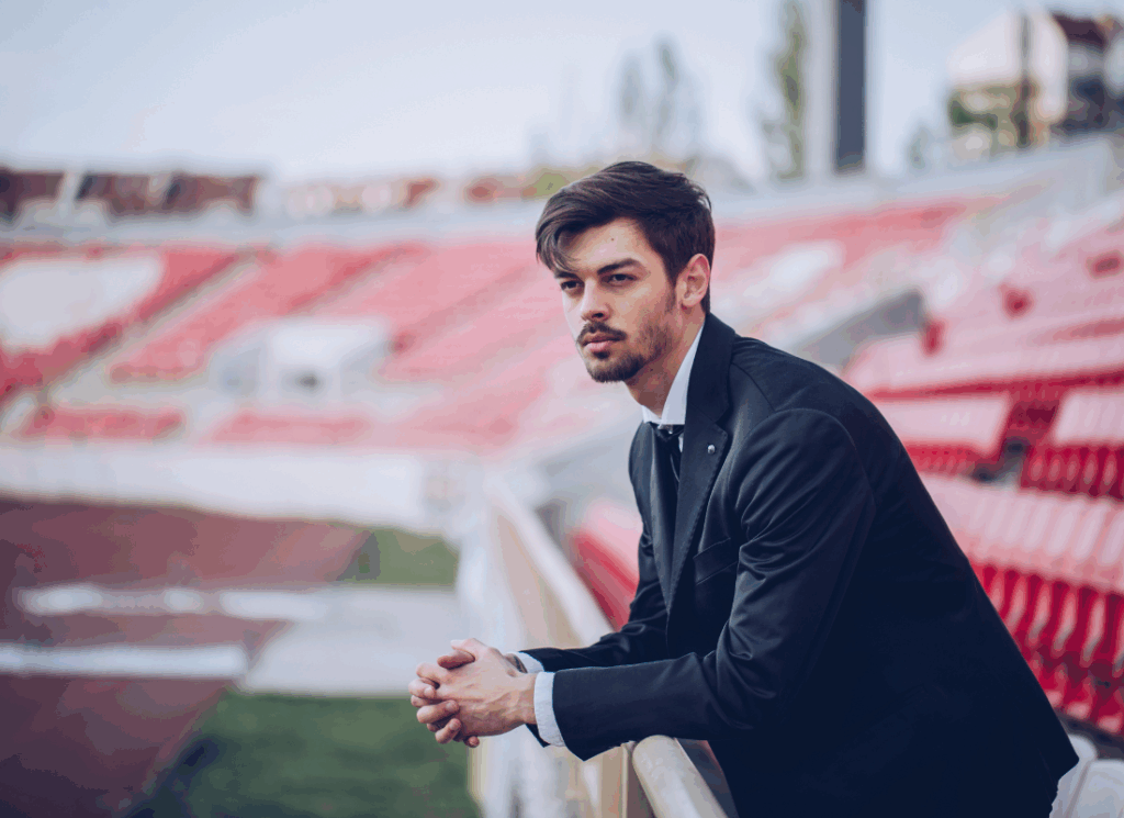 A young man in a black suit leans on a stadium railing, gazing thoughtfully over the field.