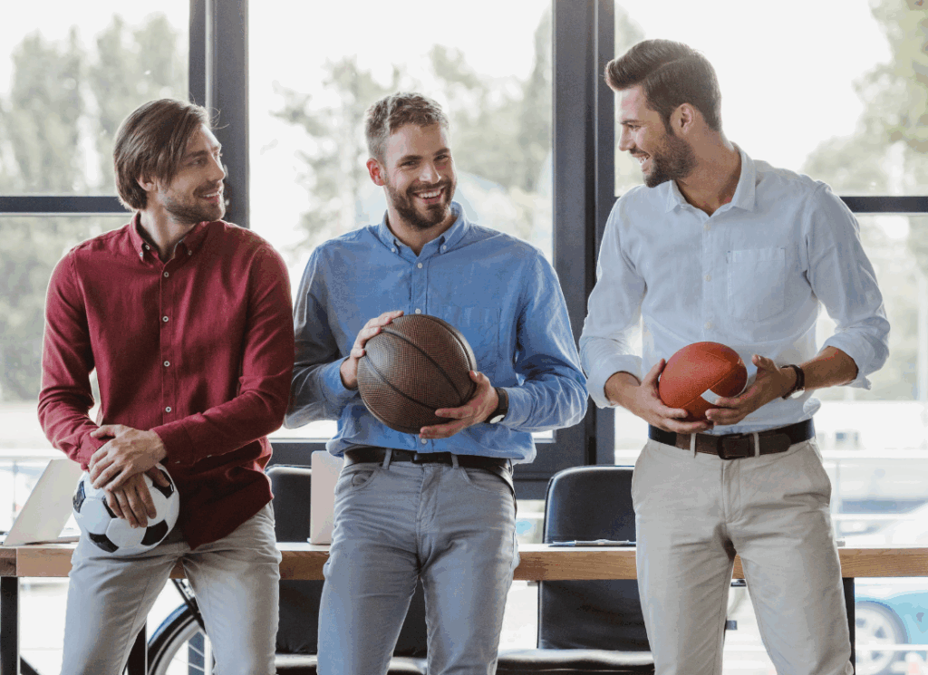 Three casually dressed businessmen stand indoors smiling and chatting, each holding a different sports ball—soccer, basketball, and football—symbolizing collaboration for starting a college athletics LLC.