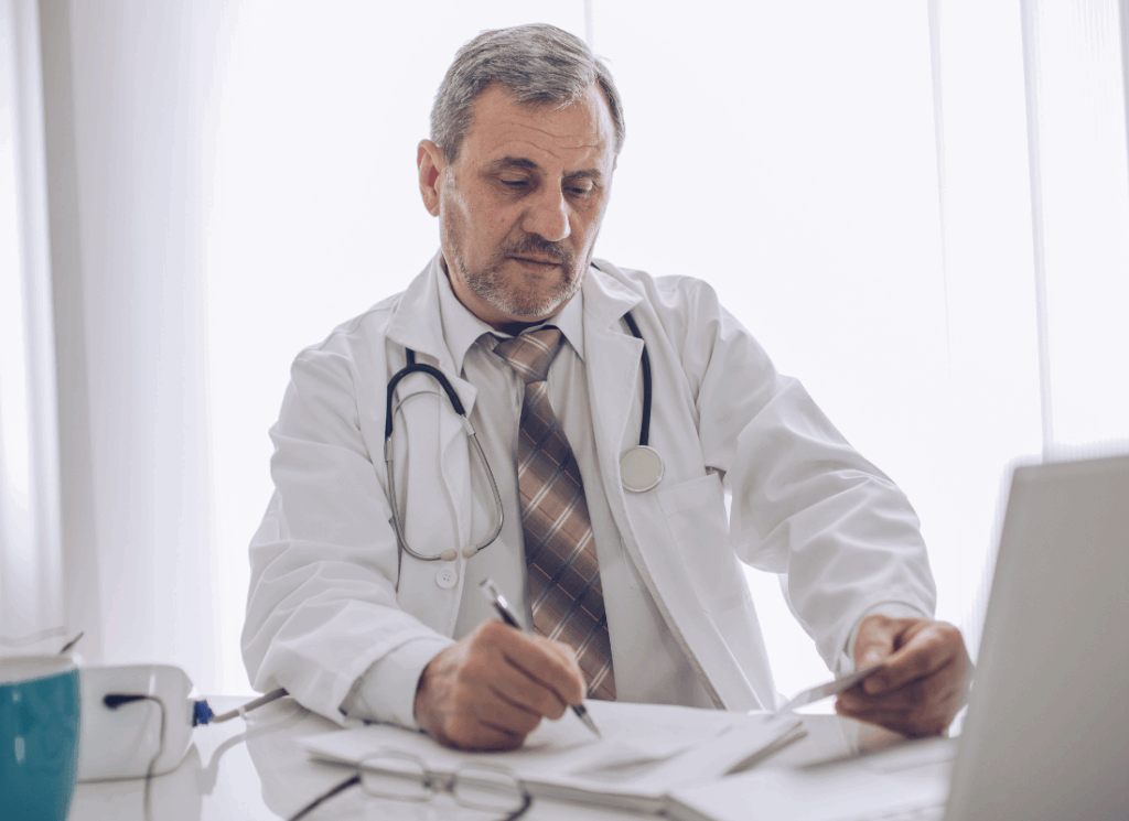 An older male doctor in a white coat with a stethoscope around his neck sits at a desk reviewing paperwork, symbolizing the importance of retirement planning for doctors.