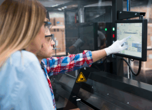 Two workers monitor and adjust settings on a touchscreen control panel in a manufacturing facility, representing the use of technology to improve manufacturing efficiency.