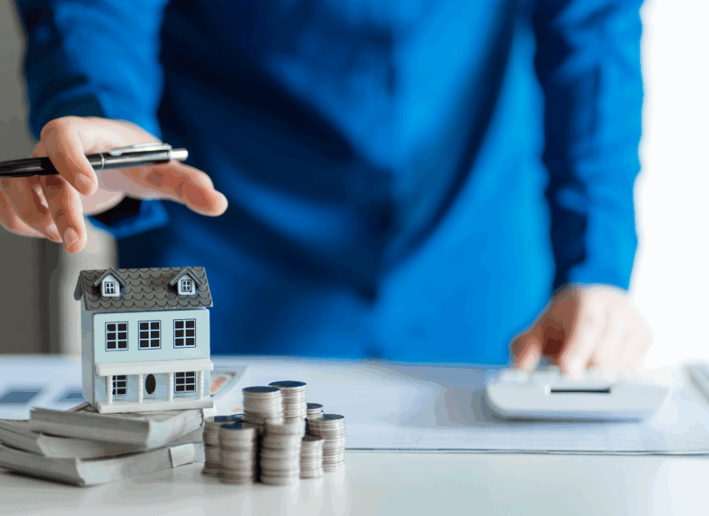 A real estate developer in a blue shirt gestures toward a miniature house model, stacks of coins, and cash, symbolizing financial planning and accounting principles for real estate development.