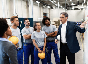 Lead Like the Workforce has Changed—Because It Has. Leadership Development. Mature businessman presenting business strategy on white board to group of industrial workers in a factory.