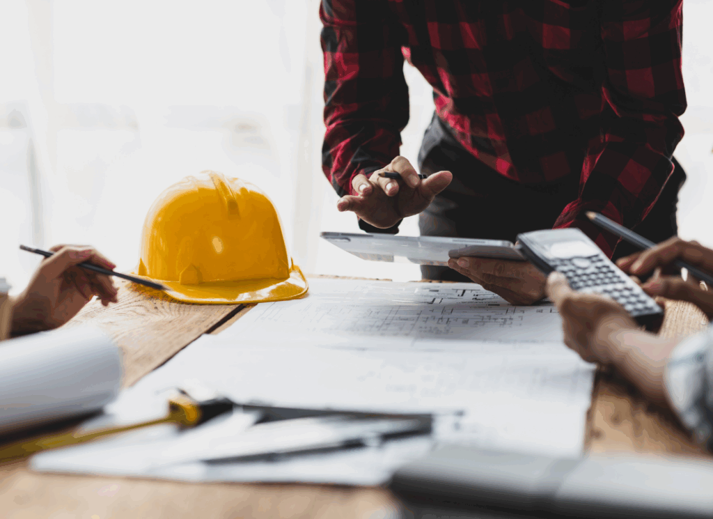 Construction accountants review blueprints and charts use calculators at a table with a yellow hard hat, symbolizing construction project accounting.