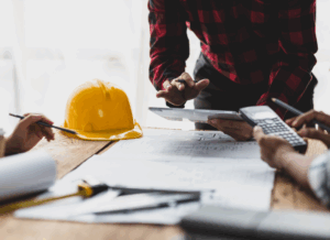 Construction accountants review blueprints and charts use calculators at a table with a yellow hard hat, symbolizing construction project accounting.