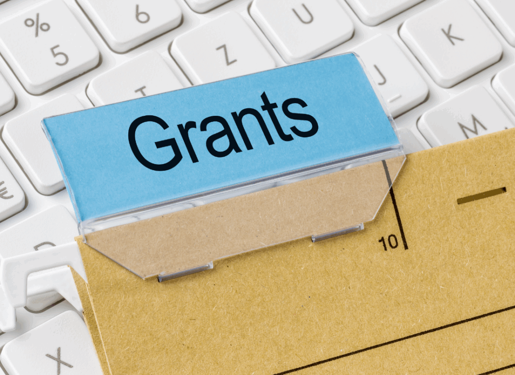 A file folder labeled “Grants” rests on a computer keyboard, symbolizing digital organization and documentation required for building a compliant grant management system.