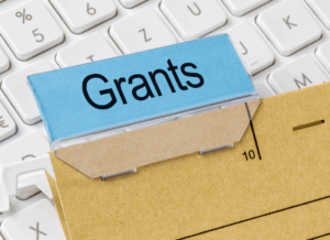 A file folder labeled “Grants” rests on a computer keyboard, symbolizing digital organization and documentation required for building a compliant grant management system.