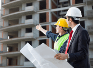 Two men wearing hard hats—one in a suit and the other in a safety vest—review blueprints and assess a building under construction, representing construction job costing and project oversight.