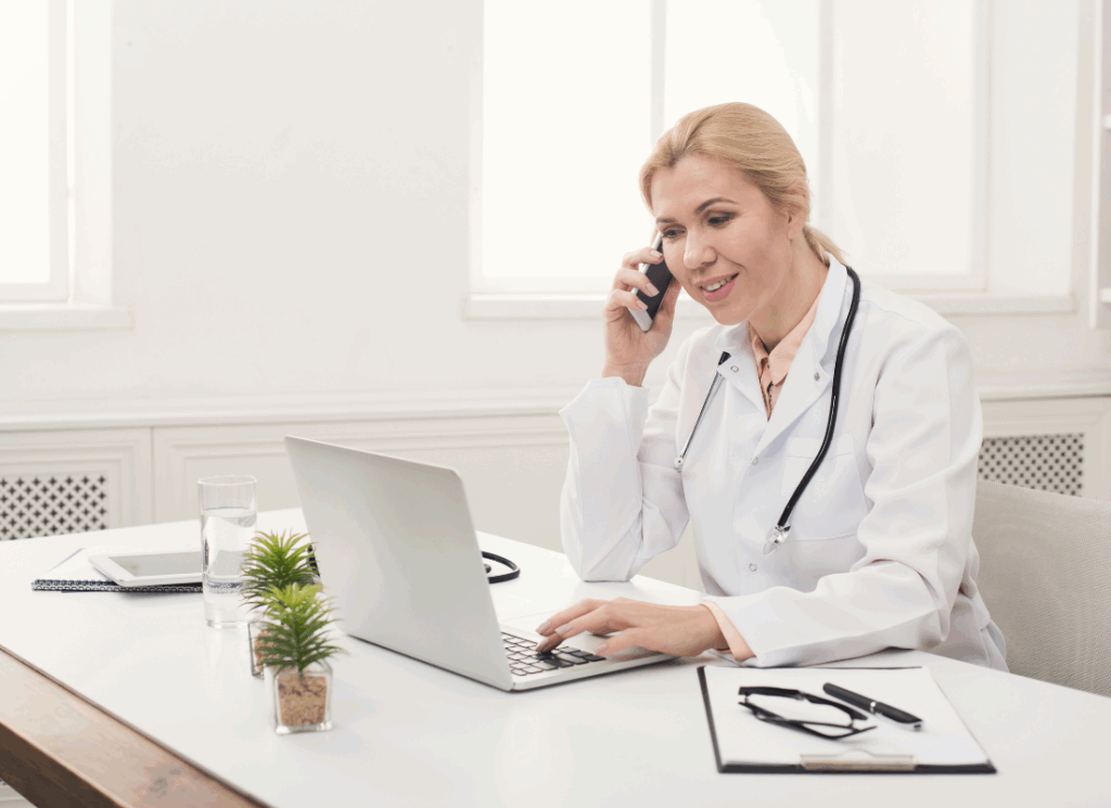 A doctor wearing a white coat and stethoscope smiles while speaking on the phone and using a laptop at a desk, representing the early stages of starting a private medical practice.