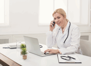 A doctor wearing a white coat and stethoscope smiles while speaking on the phone and using a laptop at a desk, representing the early stages of starting a private medical practice.