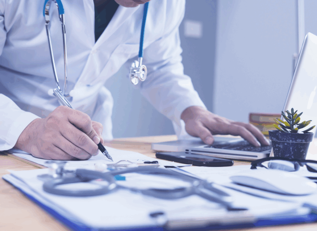 A doctor in a white coat writes notes on a clipboard while using a laptop, with medical tools and documents on the desk, representing medical practice ownership and profit allocation planning.