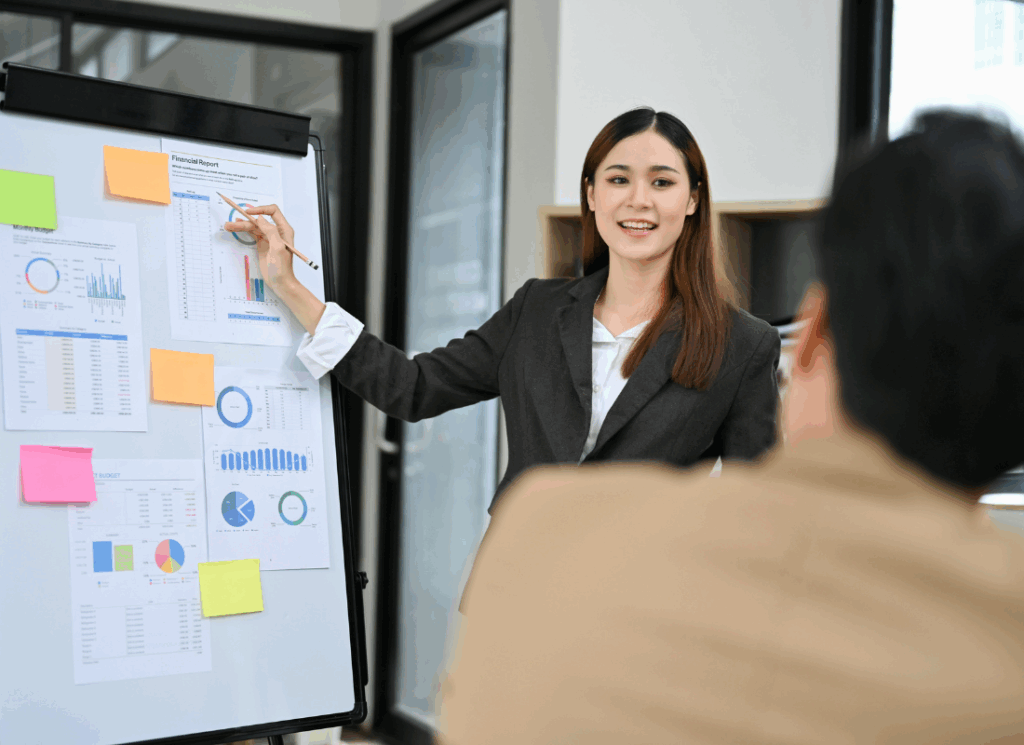 A nonprofit accounting professional in business attire presents financial charts on a whiteboard to a colleague, illustrating outsourced nonprofit accounting training.
