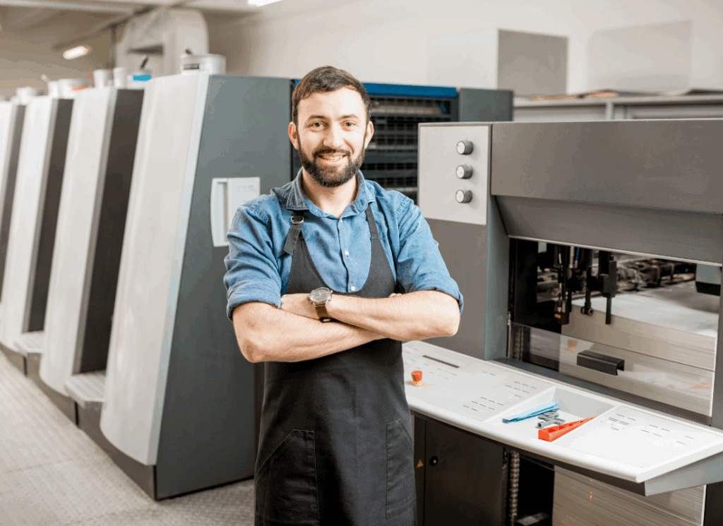 A smiling factory worker stands with arms crossed in front of industrial equipment, representing U.S. manufacturers benefiting from OBBBA and manufacturing tax reforms.
