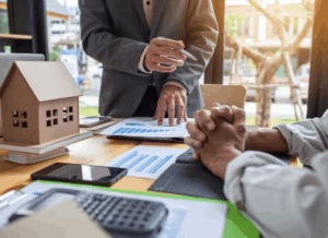 Two real estate professionals review financial charts at a desk with a model house, calculator, and documents, representing a discussion about property management accounting strategies.
