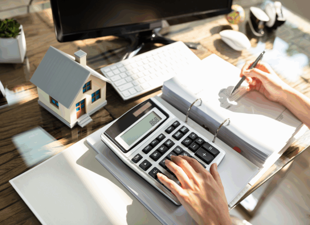 A real estate accountant uses a calculator and reviews financial documents at a desk with a miniature house model, symbolizing real estate outsourced accounting and financial planning.