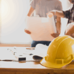 A yellow construction hard hat sits on a desk with blueprints and tools, while two construction professionals review plans related to construction forecasting.