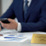 A person in a business suit reviews financial charts while holding a smartphone, with a nameplate reading “Chief Financial Officer,” symbolizing modern CFO skills in higher education.