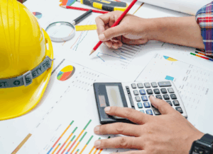 A construction accountant calculates financial data with a pencil and calculator on top of construction-related charts and graphs, next to a yellow hard hat, symbolizing construction bookkeeping.