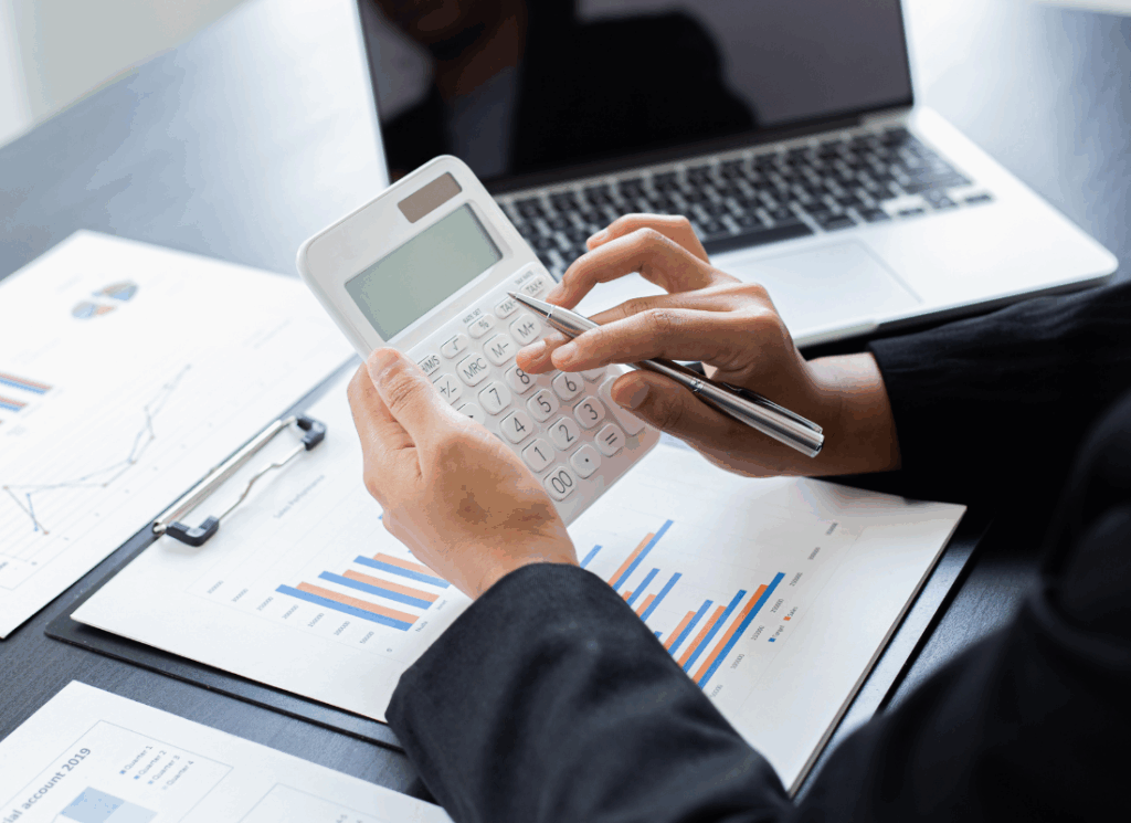 A tax accountant in business attire uses a calculator while reviewing financial charts and graphs on paper, with a laptop nearby.