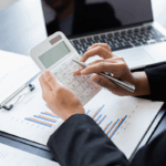 A tax accountant in business attire uses a calculator while reviewing financial charts and graphs on paper, with a laptop nearby.