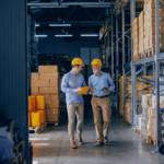Two workers in a warehouse wearing hard hats discuss documents while walking between shelves stocked with boxes—representing manufacturing company growth and operational planning.