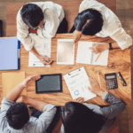 Overhead view of four nonprofit professionals gathered around a wooden table reviewing charts, tablets, and financial documents while discussing types of audits for nonprofits.