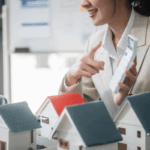 An outsourced real estate accountant gestures while holding a calculator, seated at a table with miniature house models.