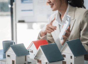 An outsourced real estate accountant gestures while holding a calculator, seated at a table with miniature house models.
