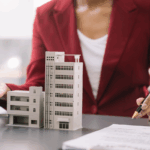 A business professional in a red blazer gestures near architectural models of commercial buildings, with documents and a pen on the table, illustrating commercial real estate accounting.