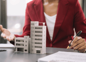 A business professional in a red blazer gestures near architectural models of commercial buildings, with documents and a pen on the table, illustrating commercial real estate accounting.