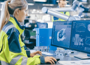 An engineer wearing a high-visibility jacket works on 3D modeling software at a dual-monitor station in a factory, illustrating continuous improvement in manufacturing processes.