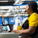 A woman wearing a headset works at a computer station inside a modern manufacturing facility, symbolizing the integration of communication and technology in the manufacturing sales process.